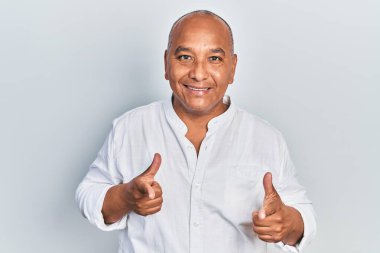 Orta yaşlı, sıradan kıyafetler giyen, mutlu ve komik yüzlü, kamerayı işaret eden Latin bir adam. iyi enerji ve titreşimler. 