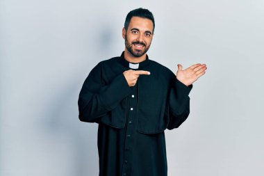 Handsome hispanic man with beard wearing catholic priest robe amazed and smiling to the camera while presenting with hand and pointing with finger. 