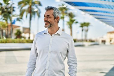 Orta yaşlı, kır saçlı, gülümseyen bir adam şehirde mutlu bir şekilde duruyor..