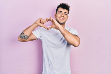 Gündelik beyaz tişört giyen, aşık gülümseyen, elleriyle kalp sembolü yapan genç İspanyol adam. romantik kavram. 