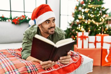 Young hispanic man reading book lying on sofa by christmas tree at home