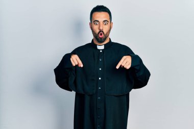 Handsome hispanic man with beard wearing catholic priest robe pointing down with fingers showing advertisement, surprised face and open mouth 