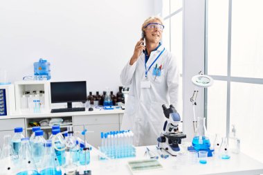 Bilim adamı üniforması giyen genç beyaz adam laboratuvarda akıllı telefondan konuşuyor.