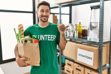 Gönüllü tişört giyen genç İspanyol adam elinde alışveriş torbalarıyla başparmağını kaldırıyor ve ağzı açık gülümsüyor. 