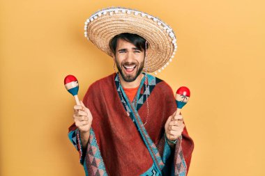 Young hispanic man wearing mexican hat holding maracas smiling and laughing hard out loud because funny crazy joke. 