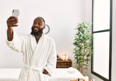 Genç Afrikalı Amerikalı gülümseyen adam güzellik merkezindeki akıllı telefondan özçekim yapıyor.