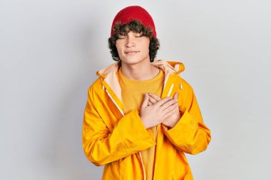 Handsome young man wearing yellow raincoat smiling with hands on chest with closed eyes and grateful gesture on face. health concept. 