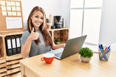 Young brunette woman working at the office with laptop doing happy thumbs up gesture with hand. approving expression looking at the camera showing success. 