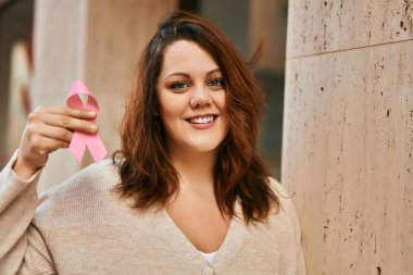 Genç İrlandalı artı cüsseli kız gülümseyerek şehirde pembe kanser kurdelesi tutuyor..