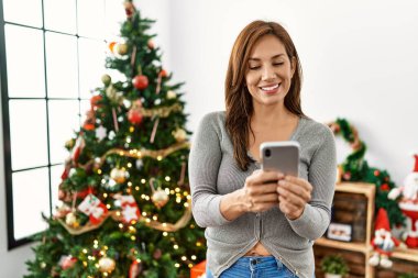 Akıllı telefon kullanan genç Latin kadın Noel ağacının yanında duruyor.