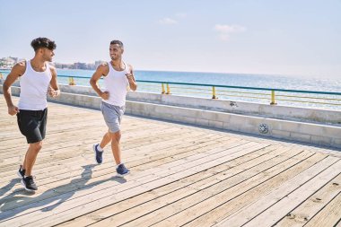İki İspanyol erkek, sportif, gülümseyen bir çift, deniz kenarında koşuyorlar.