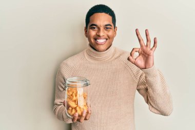 Genç, yakışıklı İspanyol adam elinde makarna kavanozuyla tamam işareti yapıyor, gülümseyerek mükemmel bir sembol oluşturuyor. 