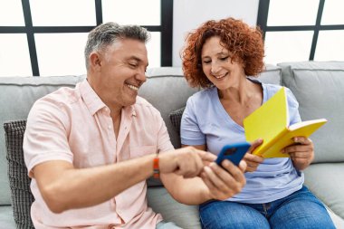Orta yaşlı erkek ve kadın çift akıllı telefon kullanıyor ve evde kanepede oturmuş kitap okuyor.