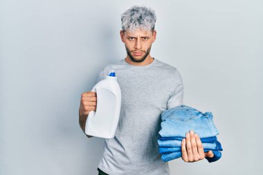 Young hispanic man with modern dyed hair holding jeans for laundry and detergent bottle depressed and worry for distress, crying angry and afraid. sad expression. 