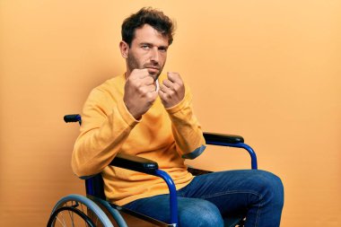 Tekerlekli sandalyede oturan sakallı yakışıklı adam. Yumruk savunma hareketiyle dövüşmeye hazır. Kızgın ve üzgün. 