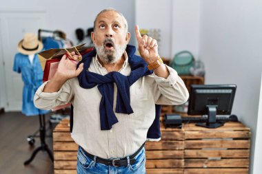 Handsome senior man holding shopping bags at boutique shop amazed and surprised looking up and pointing with fingers and raised arms. 