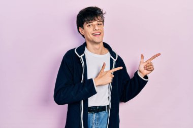 Yakışıklı hippi genç adam günlük beyaz tişört ve sweatshirt giyiyor gülümsüyor ve kameraya bakıyor iki eliyle ve parmaklarıyla yan tarafa bakıyor.. 