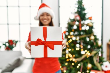 Genç Latin kadın elinde hediyeyle Noel ağacının yanında duruyor.