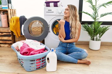 Young beautiful woman doing laundry sitting by wicker basket smiling with happy face looking and pointing to the side with thumb up. 
