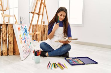 Sanat stüdyosundaki not defterine şüphe ifadesi çizen genç Latin kadın.