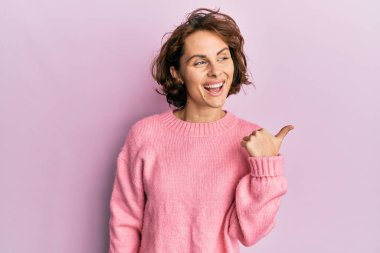 Kışlık süveter giyen genç esmer kadın baş parmağını yana kaldırmış, ağzı açık gülümsüyor. 