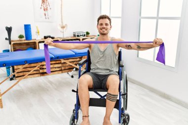 Klinikte tekerlekli sandalyede oturan elastik bant kullanarak rehabilitasyon yapan genç bir İspanyol.