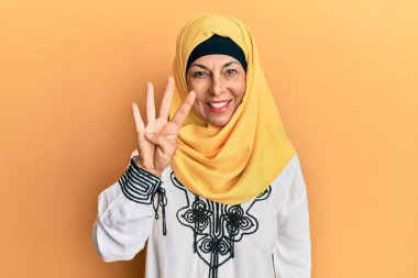 Middle age hispanic woman wearing traditional islamic hijab scarf showing and pointing up with fingers number four while smiling confident and happy. 