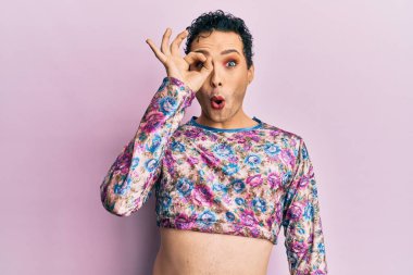 Handsome man wearing make up wearing fashion clothes doing ok gesture shocked with surprised face, eye looking through fingers. unbelieving expression. 