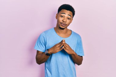 Genç Afro-Amerikalı adam, günlük mavi tişörtler giyip ellerini birleştirip gülümseyerek rahat ve neşeli bir şekilde gülümsedi. Başarı ve iyimserlik 