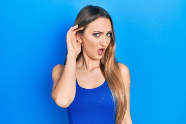 Young blonde girl doing listen gesture with hand on ear in shock face, looking skeptical and sarcastic, surprised with open mouth 
