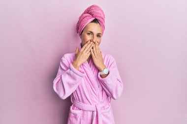 Young blonde woman wearing shower towel cap and bathrobe laughing and embarrassed giggle covering mouth with hands, gossip and scandal concept 