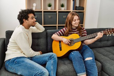 Mutlu gülümseyen genç çift evdeki kanepede oturup klasik gitar çalıyor..