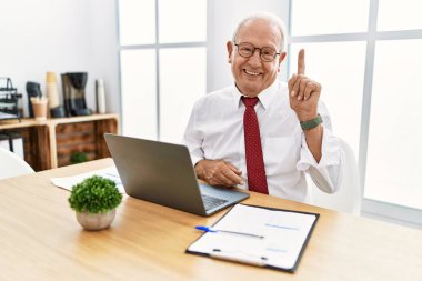 Senior man working at the office using computer laptop showing and pointing up with finger number one while smiling confident and happy. 