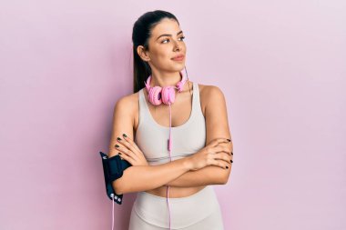 Young hispanic woman wearing gym clothes and using headphones looking to the side with arms crossed convinced and confident 