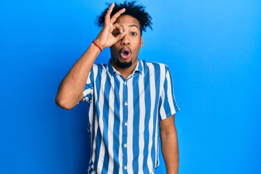 Young african american man with beard wearing casual striped shirt doing ok gesture shocked with surprised face, eye looking through fingers. unbelieving expression. 
