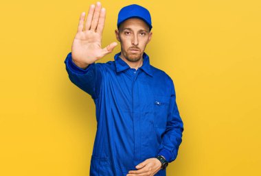Bald man with beard wearing builder jumpsuit uniform doing stop sing with palm of the hand. warning expression with negative and serious gesture on the face. 