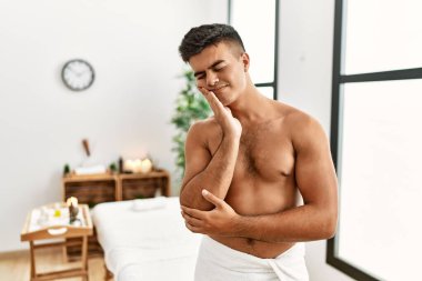 Young hispanic man standing shirtless at spa center thinking looking tired and bored with depression problems with crossed arms. 