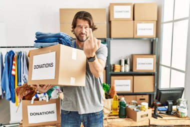 Handsome middle age man holding donations box for charity at volunteer stand showing middle finger, impolite and rude fuck off expression 
