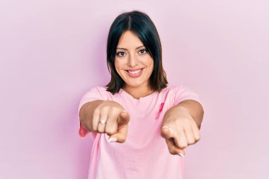 Tişörtünde pembe kanser kurdelesi olan genç İspanyol kadın parmaklarıyla seni ve kamerayı işaret ediyor. 