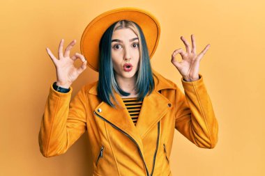Sarı şapka ve deri ceket giyen genç modern bir kız. Şaşırmış ve şok olmuş görünüyor. Onay sembolünü parmaklarıyla yapıyor. çılgın ifade 