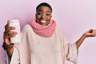 Young african american woman wearing winter scarf and drinking hot take away coffee screaming proud, celebrating victory and success very excited with raised arm 