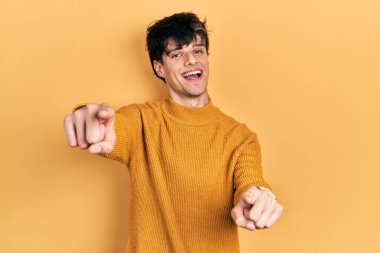 Yakışıklı hippi genç adam gündelik sarı süveter giyip seni ve kamerayı işaret ediyor parmaklarıyla, gülümsüyor pozitif ve neşeli. 