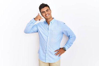 Young hispanic man wearing business shirt standing over isolated background smiling doing phone gesture with hand and fingers like talking on the telephone. communicating concepts. 