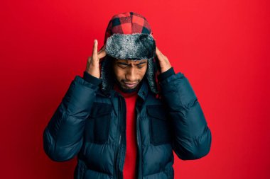 Young african american man with beard wearing winter hat and coat with hand on head for pain in head because stress. suffering migraine. 