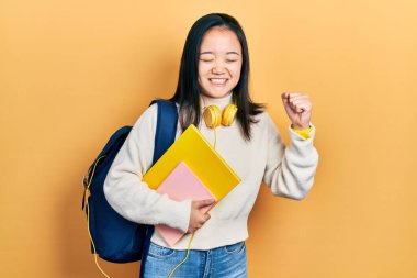 Young chinese girl holding student backpack and books very happy and excited doing winner gesture with arms raised, smiling and screaming for success. celebration concept. 