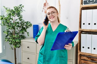 Fsiolog üniforması giyen genç kızıl saçlı kadın klinikteki akıllı telefondan konuşuyor.