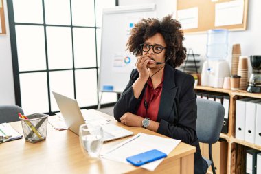 African american woman with afro hair working at the office wearing operator headset looking stressed and nervous with hands on mouth biting nails. anxiety problem. 
