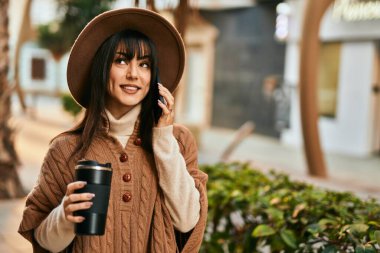 Kışlık şapkalı esmer kadın telefonda konuşuyor ve şehirde bir fincan kahve içiyor.