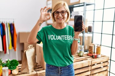 Middle age blonde woman working wearing volunteer t shirt showing smartphone screen doing ok sign with fingers, smiling friendly gesturing excellent symbol 