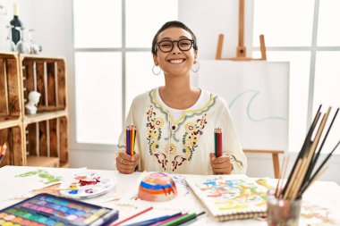 Resim stüdyosunda elinde renkli kalemlerle gülümseyen İspanyol bir kadın.